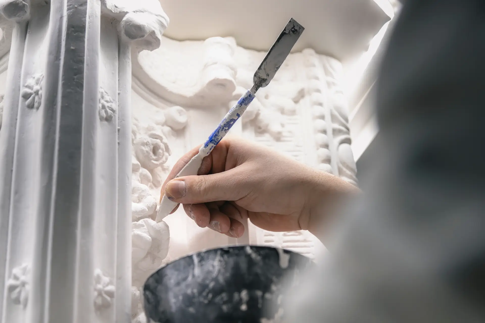 Hand eines Stukkateurs, der mit einem kleinen Spachtel feine Details an einer verzierten, weißen Stuckatur ausbessert.