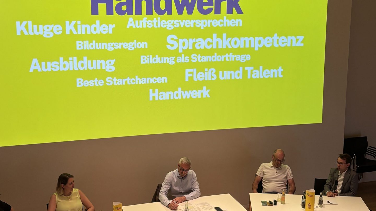 u.a. Yannic Schmitt sitzt im Podium an der RWTH vor einer Beamerfläche auf der Worte wie Handwerk und Kluge Kinder angebeamt wird.